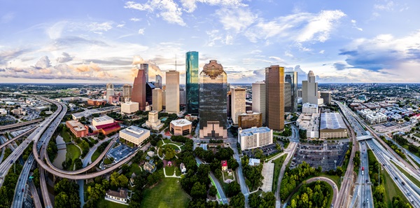 Aerial view of downtown Houston, Texas, with skyscrapers, highways, and green spaces under a partly cloudy sky.